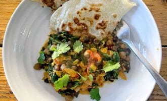 A white plate with chickpea and spinach curry garnished with coriander, served with two pieces of flatbread. A spoon rests on the plate, which is set on a wooden table.