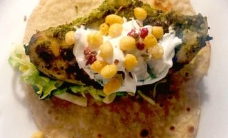 A wholemeal tortilla topped with grilled chicken, shredded lettuce, a dollop of soured cream, crispy round snacks, and red seasoning, served on a white plate.