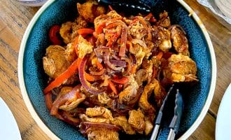 A bowl of cooked chicken strips with red onions and red peppers, served in a blue bowl with tongs. Grated cheese and tortillas are visible nearby on a wooden table.
