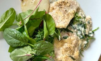 A white plate with creamy chicken and spinach, a halved boiled potato, and a fresh green salad with leafy greens.