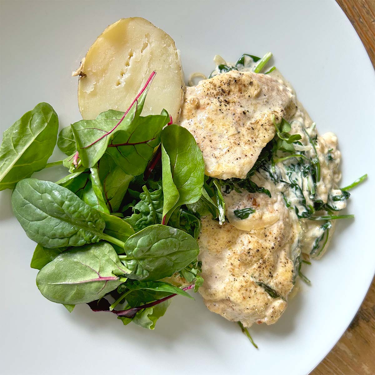 A white plate with creamy chicken and spinach, a halved boiled potato, and a fresh green salad with leafy greens.