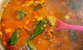 A pot of red, spicy stew with chunks of meat and whole green peppers, being stirred with a pink spoon. Steam rises from the hot, simmering dish.