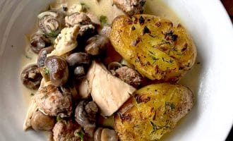A white bowl contains creamy chicken and mushroom stew with herbs, meatballs, and two crispy, golden roasted smashed potatoes on the side.
