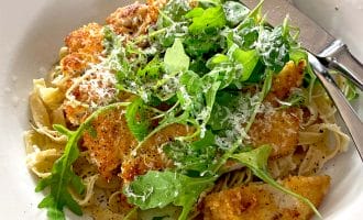A white plate with pasta topped with crispy breaded chicken, fresh rocket, and grated cheese, with a fork and knife resting on the plate.