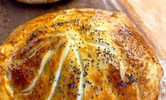Golden-brown, round pastry topped with poppy seeds and decorative scored lines, resting on a baking tray. Another similar pastry is visible in the background.