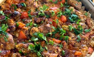 A frying pan filled with arroz con pollo, showing seasoned chicken, rice, tomatoes, red peppers, and fresh parsley on top. The dish looks freshly cooked and colourful.