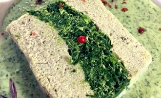 A rectangular slice of pâté with a green spinach centre is served on a creamy green herb sauce, garnished with pink peppercorns and small purple microgreens.