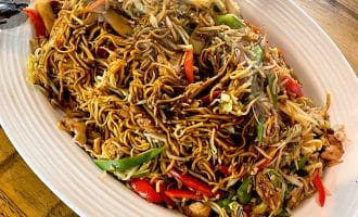 A large oval platter of stir-fried noodles with vegetables, chicken, and red and green peppers is placed on a wooden table, with serving tongs on each end of the dish. Steam rises from the hot food.