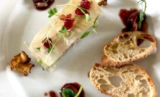 A gourmet dish presented on a white plate with two slices of crusty bread, a rectangular portion of food topped with microgreens and sauce, garnished with mushrooms and olives, and artistic sauce drizzles.