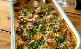 A baking dish filled with creamy meatballs garnished with chopped parsley, placed on a wooden table next to a glass of white wine and some drinking glasses.