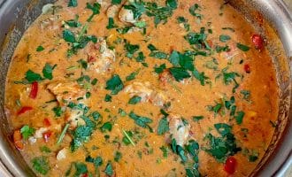 A metal pan filled with creamy, orange-coloured chicken stew, garnished with chopped fresh parsley and pieces of red pepper, sitting on a wooden surface.