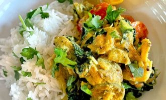 A white plate with steamed white rice, creamy yellow curry with chicken, spinach, tomatoes, and fresh coriander, garnished with a lemon wedge.
