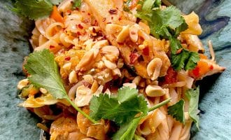 A close-up of a vibrant noodle dish topped with chopped peanuts, fresh coriander, red chilli flakes, and pieces of chicken, served in a green bowl.