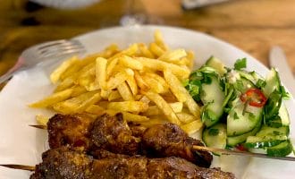A plate with two grilled meat kebabs, a serving of golden chips, and a fresh cucumber salad garnished with herbs and red chilli slices. A fork and knife rest on the table beside the plate.