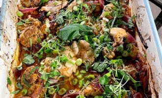 A baked dish filled with roasted chicken pieces, red onions, and herbs like coriander and mint, garnished with sliced spring onions and microgreens. The food is in a white rectangular baking dish on a hob.