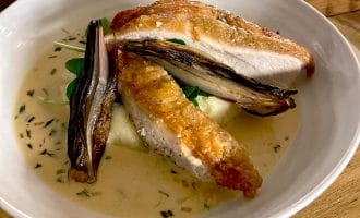 A white bowl with roasted chicken breast on creamy mashed potato, topped with grilled vegetables and a light herb sauce, served on a wooden table.