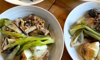Four white bowls filled with baked potatoes, grilled spring onions, mushrooms, and creamy sauce are arranged on a wooden surface. The dish appears hearty and rustic, featuring roasted and sautéed vegetables.