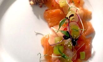 Thin slices of raw salmon arranged on a white plate, garnished with microgreens, radish slices, cucumber, and a small pile of crumbled topping on the side.