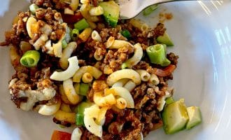 A white plate holds a serving of macaroni pasta mixed with minced beef, diced spring onions, avocado, tomato, and melted cheese. A fork rests on the edge of the plate.