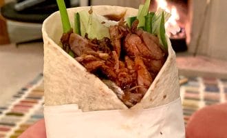 A hand with red nail varnish holds a wrap filled with shredded meat, lettuce, and cucumber. A fireplace burns in the background, creating a cosy indoor atmosphere.