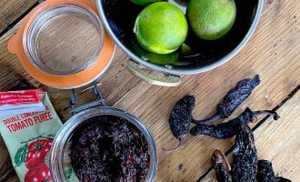 A bowl of three limes, a glass jar of dark paste, dried chillies, a tube of tomato purée, and an empty jar are arranged on a wooden surface.