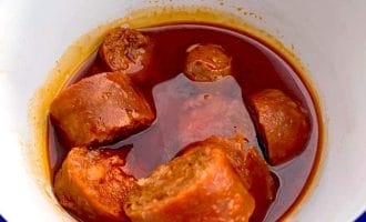 A white bowl with a blue rim contains several pieces of sausage in a rich, oily red sauce, likely a spicy or tomato-based stew.