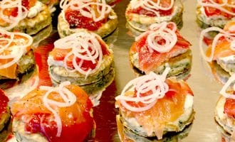 Rows of small, round appetisers on a metal tray, topped with cream, slices of smoked salmon, and thinly sliced onions. The appetisers are neatly arranged and garnished for serving.