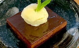 A square slice of chocolate cake topped with a scoop of vanilla ice cream and a fresh mint leaf, served on a dark ceramic plate with a rustic wooden background.