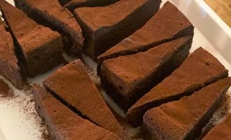 Slices of rich, chocolate cake dusted with cocoa powder are arranged neatly on a white rectangular plate on a wooden table.
