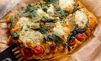 A rectangular baked frittata with spinach, cherry tomatoes, and melted cheese sits on baking parchment. A spatula is lifting a slice, and a knife rests beside it. The dish appears golden and freshly baked.
