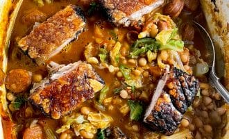 A pot filled with crispy roast pork belly pieces on top of a hearty stew with beans, sausage slices, chopped cabbage, and a rich, tomato-based broth. A spoon rests in the stew.