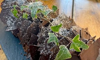 A chocolate yule log cake decorated with ivy leaves and dusted with icing sugar sits on a black slate platter atop a wooden table.