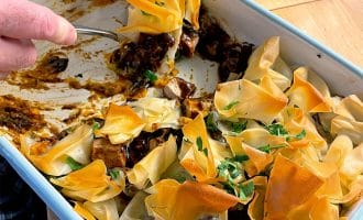 Hands serving a portion of a baked dish with crispy, golden filo pastry on top, garnished with chopped parsley, from a rectangular baking dish on a wooden table. Some filling and pastry remain in the dish.