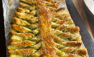 A baked puff pastry shaped like a Christmas tree, with twisted branches and sprinkled with sesame seeds, sits on baking parchment atop a wooden surface.