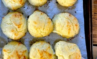 Nine golden-brown, fluffy baked scones are arranged on baking parchment in a baking tray. One scone is being lifted with a spatula, showing its light, airy texture.