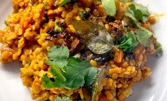A serving of orange lentil dahl garnished with fresh coriander leaves and bay leaves, plated on a white dish. The dahl appears creamy with visible spices and herbs mixed in.