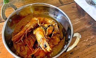 A metal bowl with ornate handles holds a prawn curry in a rich, reddish sauce. The bowl sits on a rustic wooden table beside a folded serviette with a fork and spoon, and some fresh herbs are visible nearby.