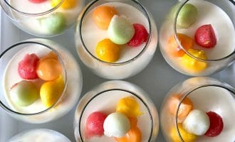 Several glasses filled with a creamy white dessert are topped with colourful melon balls in shades of orange, yellow, red, and green, arranged neatly on a white surface.