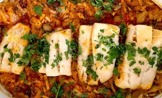 A baked dish featuring four pieces of white fish fillet on a bed of seasoned orzo pasta, topped with chopped fresh herbs, all served in a white oval baking dish.