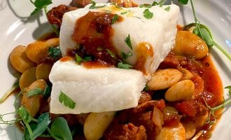 A dish featuring a piece of white fish atop a stew of large white beans, tomato-based sauce, and herbs, garnished with fresh greens on a white plate.