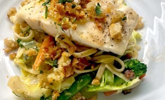 A piece of white fish topped with crispy breadcrumbs sits on a bed of mixed vegetables and pasta, served on a white plate.