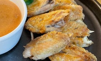 Crispy baked chicken wings arranged on a black plate, served with a pot of orange dipping sauce and a garnish of fresh coriander.