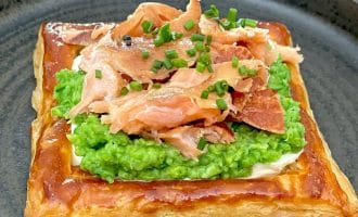 A square puff pastry topped with a layer of mashed peas, flaked smoked salmon, and chopped chives, served on a dark round plate.