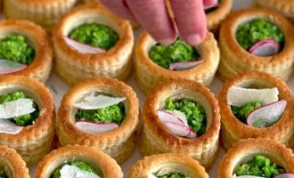 A hand garnishes round puff pastry shells filled with garden peas, topped with radish slices and cheese shavings, arranged neatly on a tray.