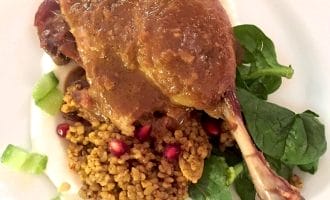 A cooked duck leg with brown sauce is served on a bed of spinach, bulgur or couscous, pomegranate seeds, and diced celery, plated on a white plate.