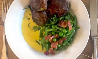 A white plate with three pieces of glazed meat, green beans and peas topped with bacon pieces, and yellow sauce, set on a wooden table with a fork and knife beside the plate.