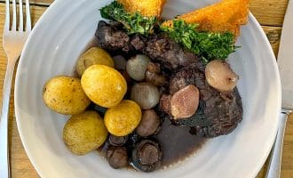 A plate with boiled baby potatoes, pearl onions, mushrooms, braised meat in dark sauce, and two triangular pieces of toast topped with chopped herbs. Knife and fork are placed on either side of the plate.