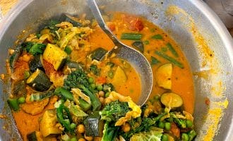 A large metal bowl filled with vibrant vegetable curry, including courgette, broccoli, peas, green beans, and chickpeas in a rich orange sauce, with a ladle resting inside. A bowl of white rice is in the background.