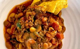A plate of chilli with beans, minced meat, tomatoes, and vegetables, topped with three yellow corn tortilla crisps on the side. The chilli appears hearty and saucy, served on a white plate.