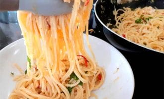 A close-up of spaghetti being served from a frying pan into a white bowl using kitchen tongs. The pasta is mixed with herbs and sliced red chilli peppers.